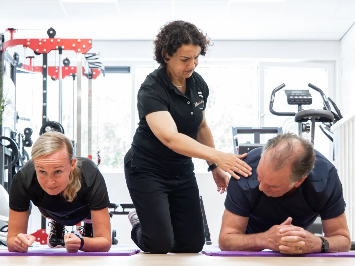 Miriam corrigeert de houding van twee klanten die naast elkaar in plankpositie op matten trainen