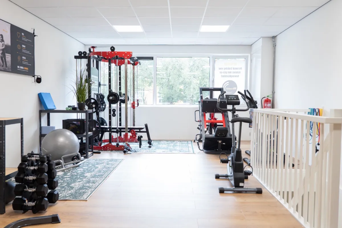 De Mpowher Huiskamer Gym in Apeldoorn: lichte ruimte met houten vloer, krachtstation, dumbbells en hometrainers