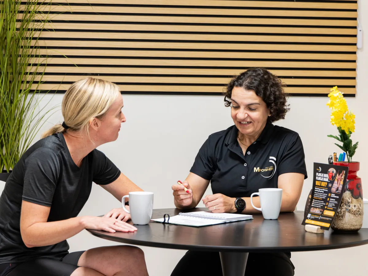 Miriam in gesprek met een klant aan de tafel in de Huiskamer Gym, allebei met een kop koffie