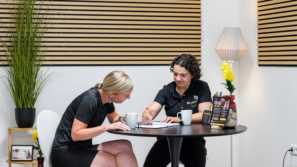 Miriam en een klant in gesprek aan de tafel in de Huiskamer Gym, met koffie en een notitieblok