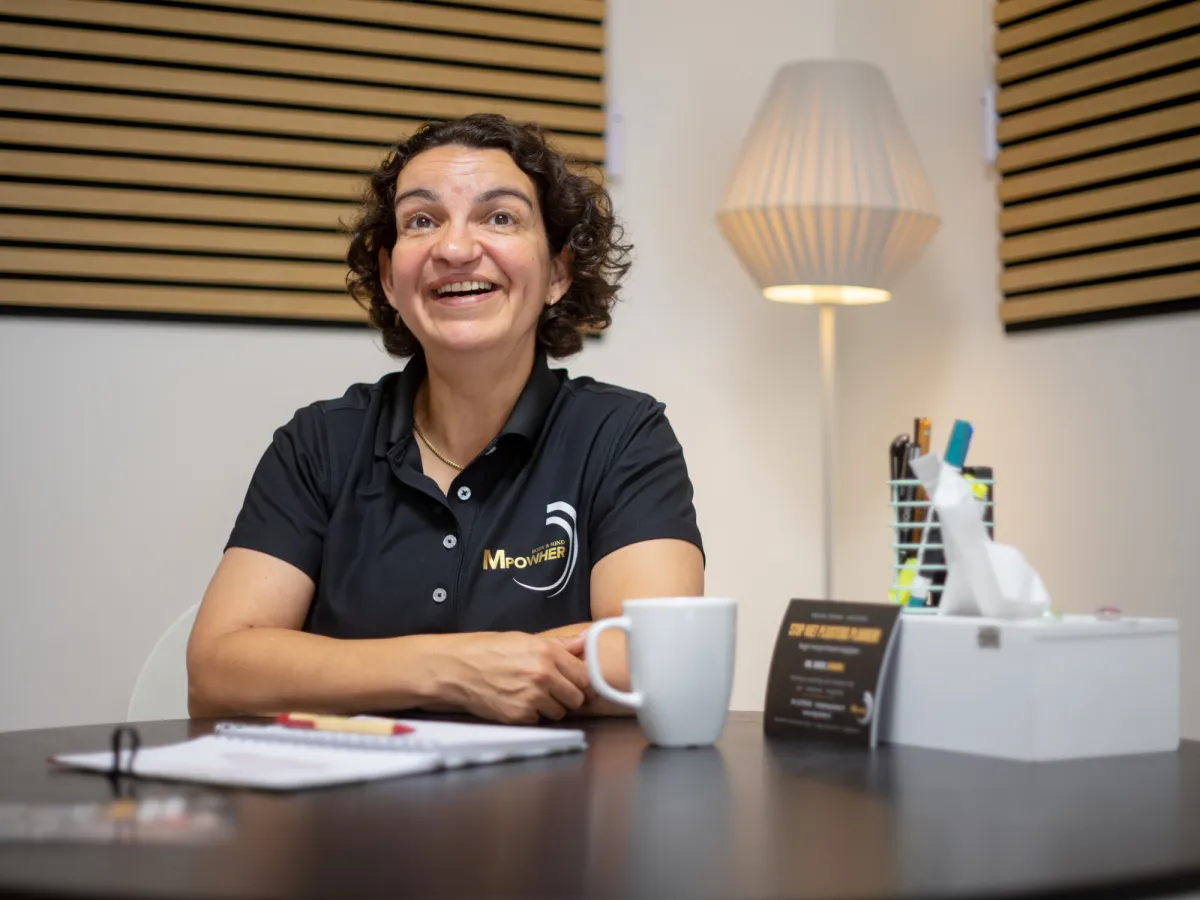 Miriam Geerdes-Gazzah lachend aan de tafel in haar Huiskamer Gym in Apeldoorn, met een kop koffie en een notitieblok